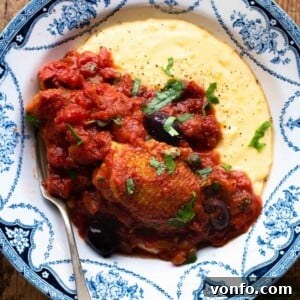 A cropped close up of chicken puttanesca with creamy polenta in a blue bowl, garnished with fresh herbs.