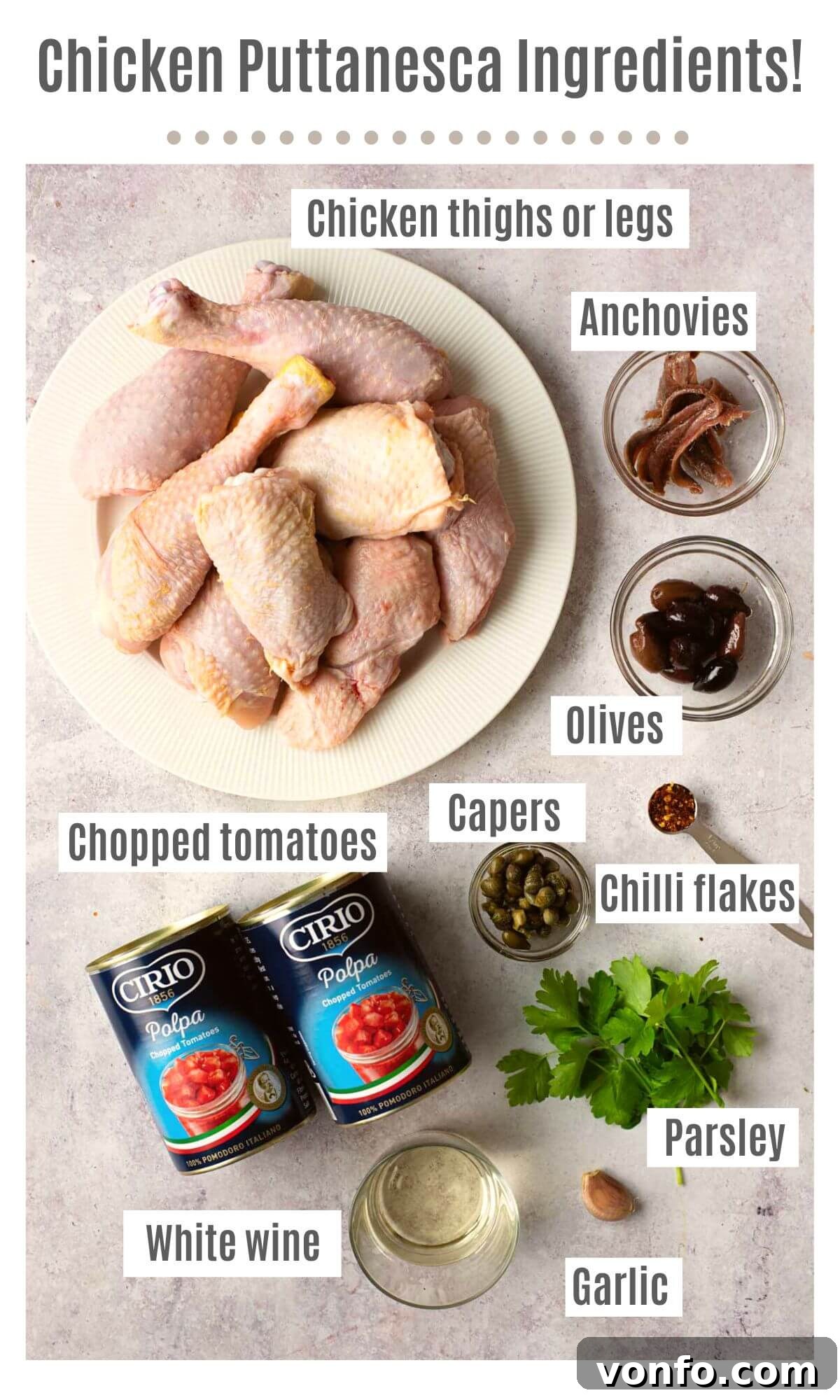 An overhead shot of labelled ingredients needed to make chicken puttanesca; chicken thighs or legs, anchovies, olives, capers, chilli flakes, chopped tomatoes, parsley, garlic and white wine, neatly arranged on a white surface.