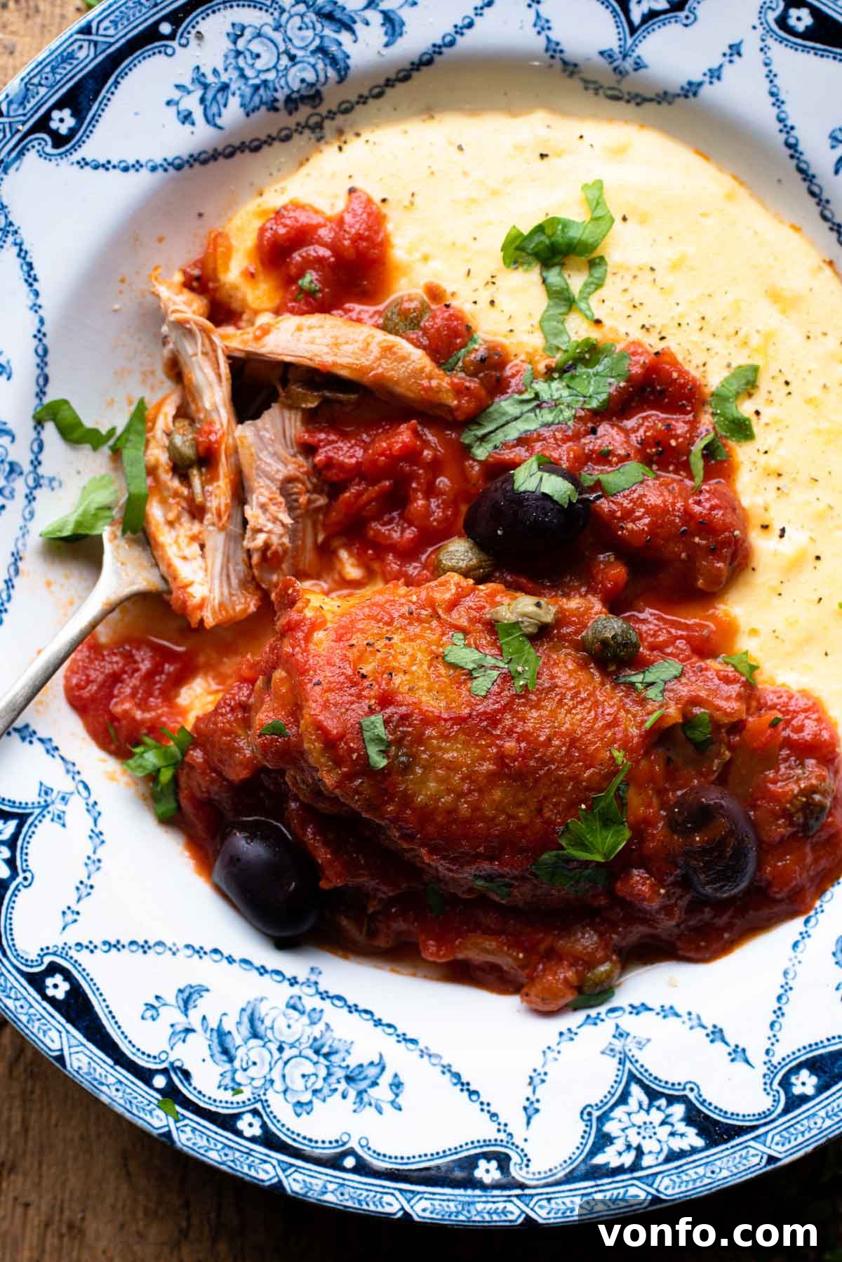 Chicken in Puttanesca sauce in a bowl with polenta sitting on a wooden surface, ready to be enjoyed as a meal.