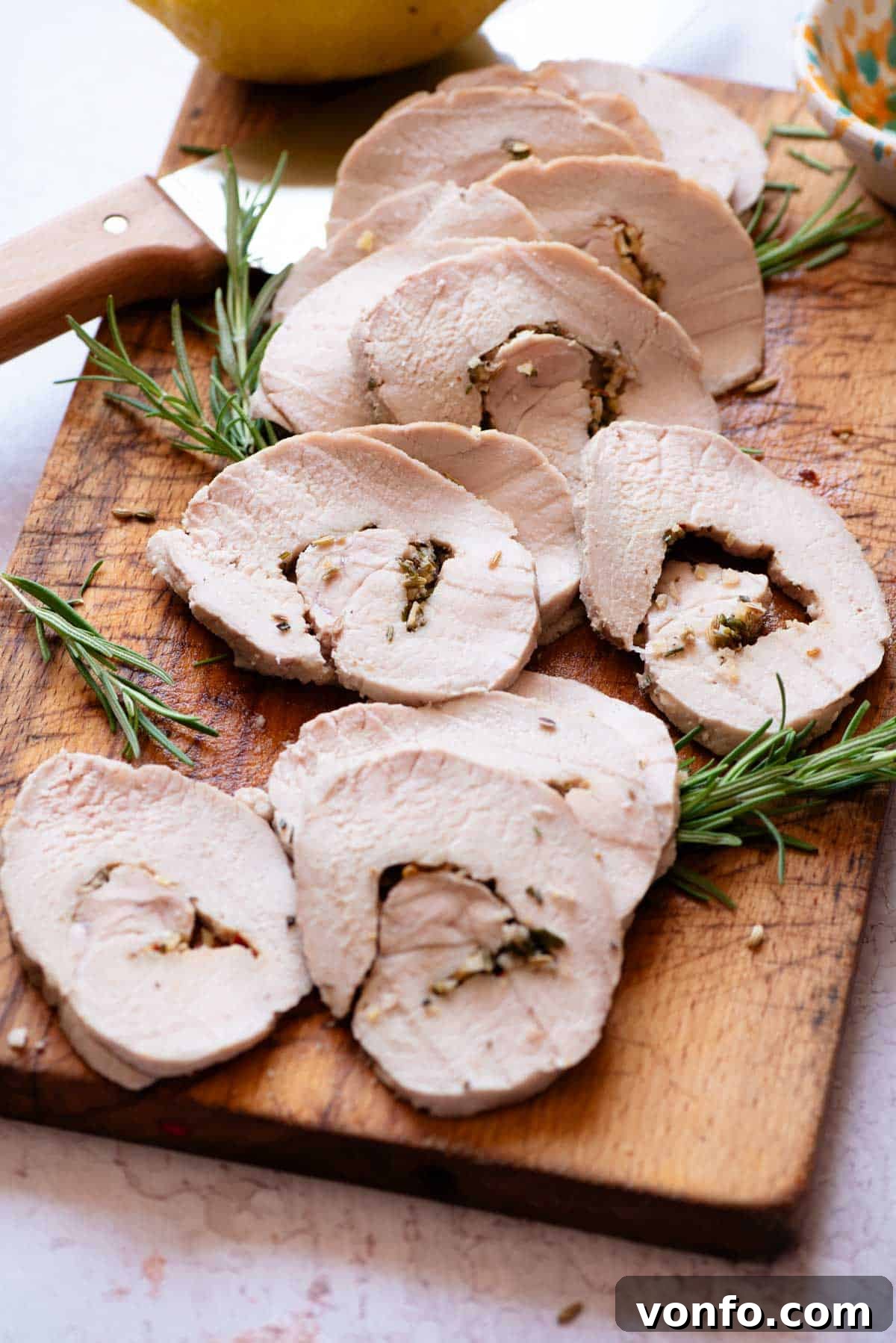 Slices of Italian roast pork loin on a wooden chopping board, garnished with fresh lemon and herbs, with a carving knife nearby.