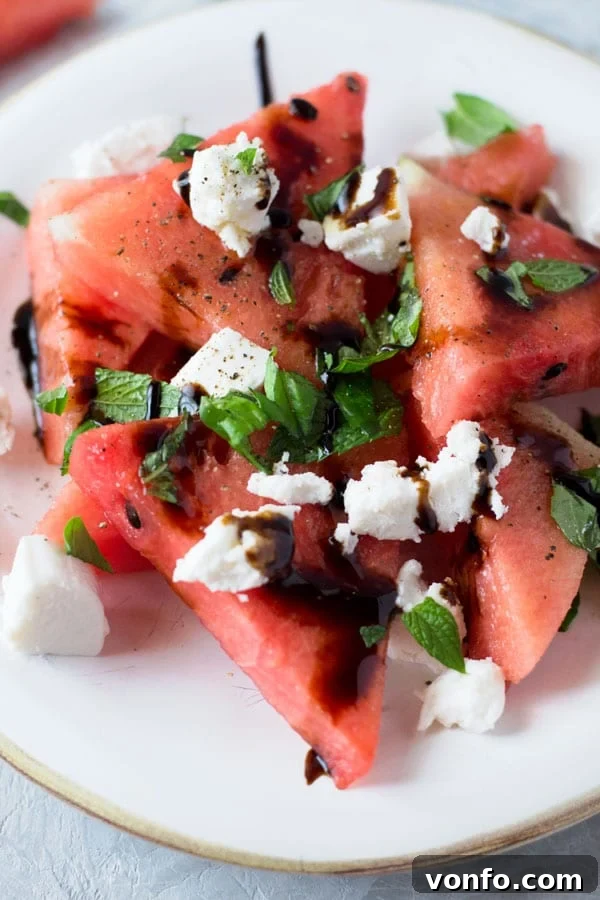 A close-up shot of a serving of watermelon basil salad, garnished with mint and goat cheese, drizzled with balsamic glaze. Healthy and delicious.