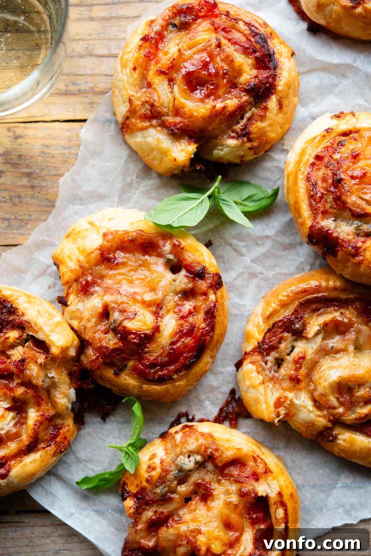 Prosciutto Gorgonzola Pizza Swirls 2 An overhead shot of golden-brown pizza pinwheels arranged beautifully on a rustic wooden board, garnished with a fresh basil leaf and a glass of red wine in the background.