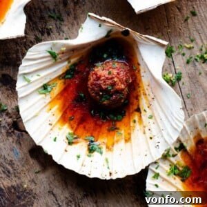 A close up of a scallop in a shell topped with vibrant 'Nduja garlic butter and a hint of fresh herbs.
