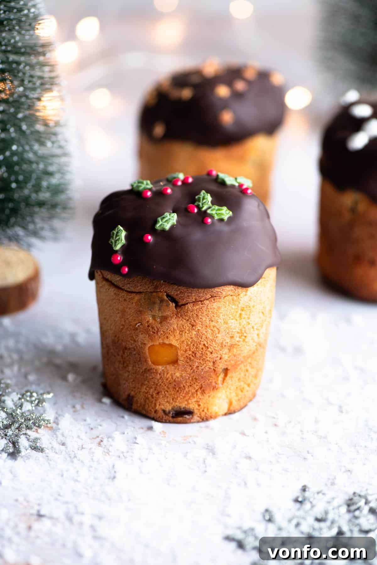 Cherry Chocolate Ice Cream Panettone Indulgence 2 A beautifully decorated mini Panettone, dipped in melted dark chocolate and adorned with festive sprinkles, sits on a snowy Christmas-themed surface, flanked by miniature Christmas trees. The focus is on the rich chocolate topping and the promise of a delightful treat inside.