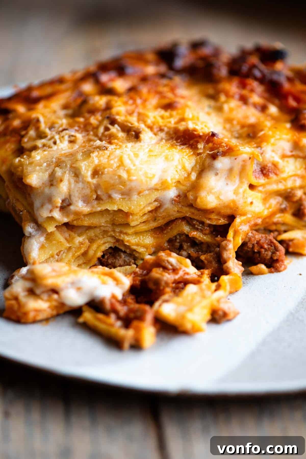 Hearty Lamb Lasagna with Pecorino Romano Béchamel 2 One slice of lamb lasagna on a plate sitting on a wooden surface, showcasing rich layers and golden-brown top.