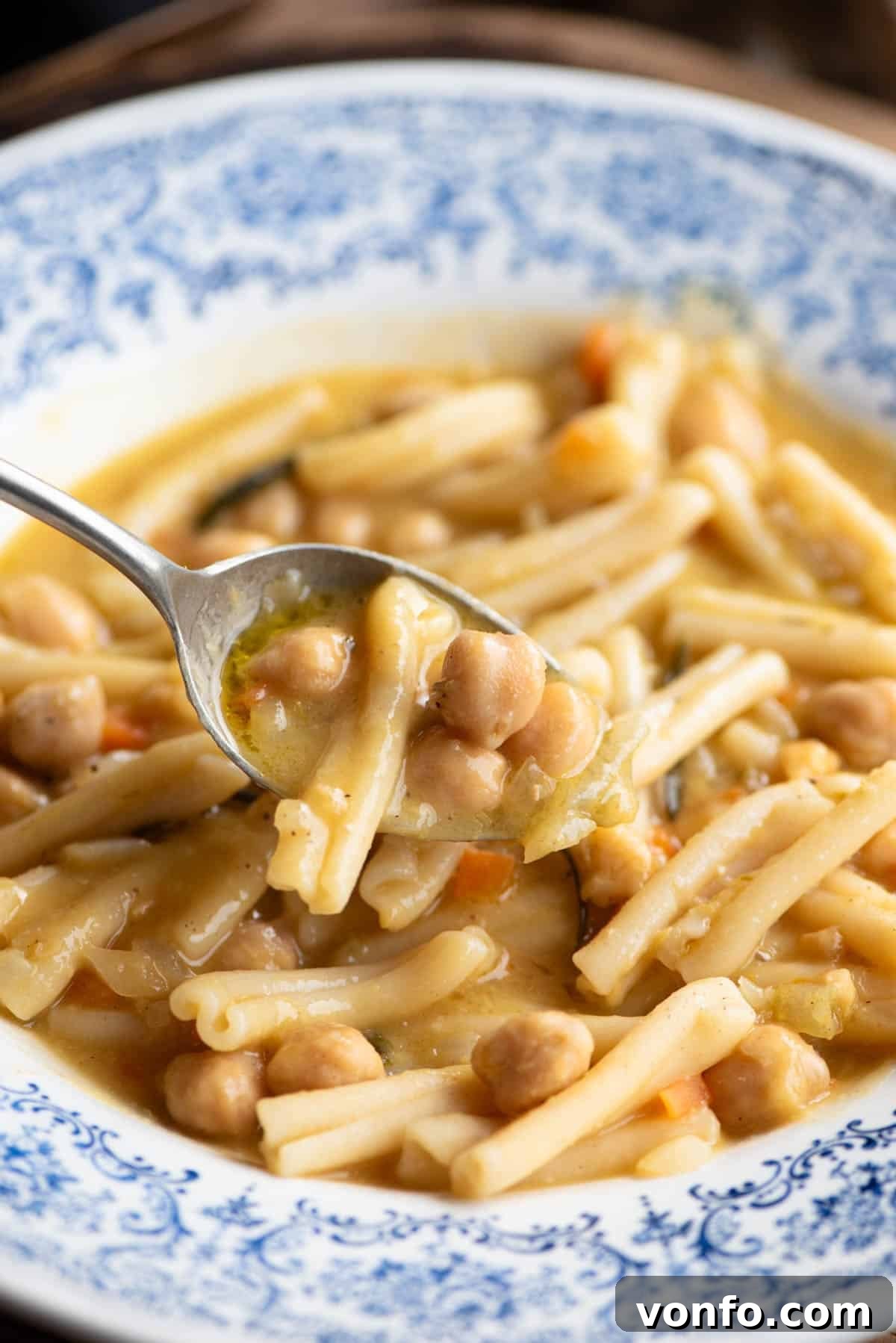 A close-up view of a spoon laden with creamy Pasta e Ceci, showing the perfect blend of pasta and chickpeas, held invitingly above a blue ceramic bowl.