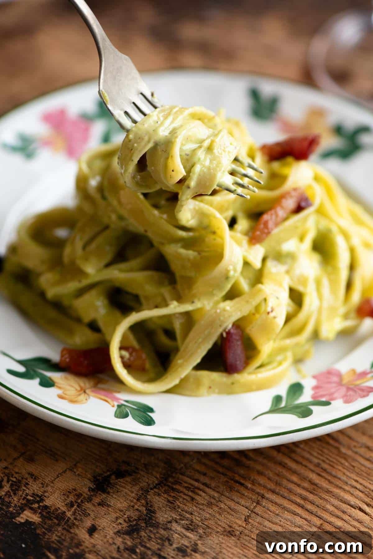 A fork elegantly lifting a generous portion of tagliatelle coated in creamy pistachio sauce from a beautiful floral patterned plate, showcasing the dish's rich texture and inviting allure, ready to be savored.