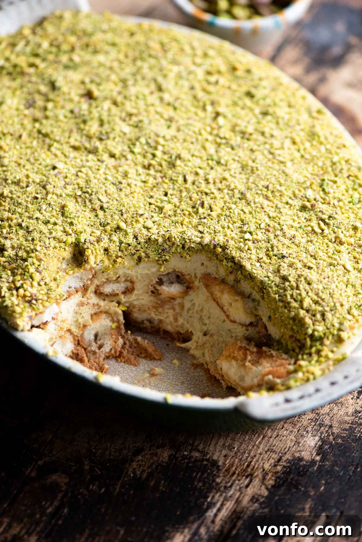 Green Gold Tiramisu 9 A side shot of a pistachio tiramisu in an oval dish with a slice removed, displaying the layers beautifully. The background is a rustic wooden table.