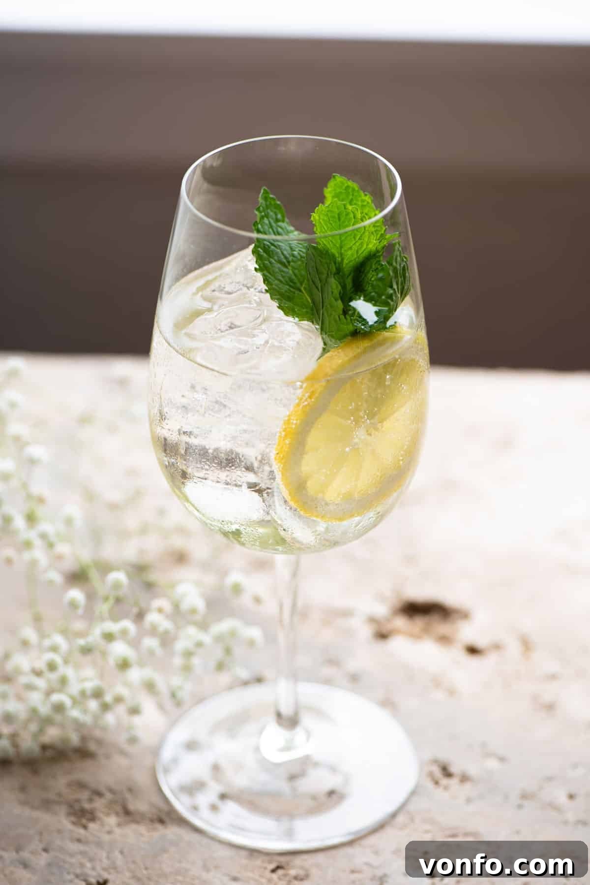 Elderbloom Sparkler 4 A Hugo Spritz cocktail, glistening with ice and garnished with a sprig of mint and lemon, rests on a textured stone worktop, perfect for a sunny day.