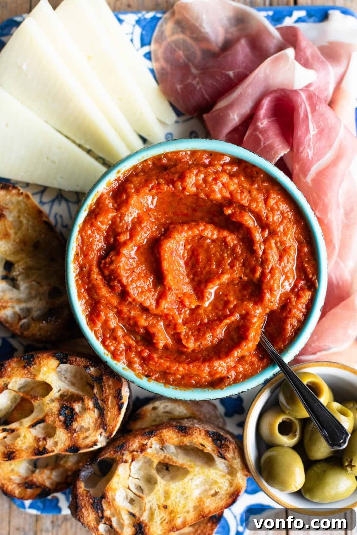 A close up of Ajvar dip in a blue bowl surrounded by cured meat, cheese, ciabatta bread and olives. This image showcases the vibrant color and texture of homemade Ajvar, perfect for antipasti.