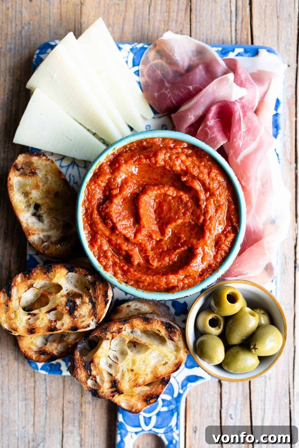 An overhead shot of Ajvar spread in a blue bowl sitting on a serving board with cured meats, cheese and olives, emphasizing its role in a rich antipasti spread.