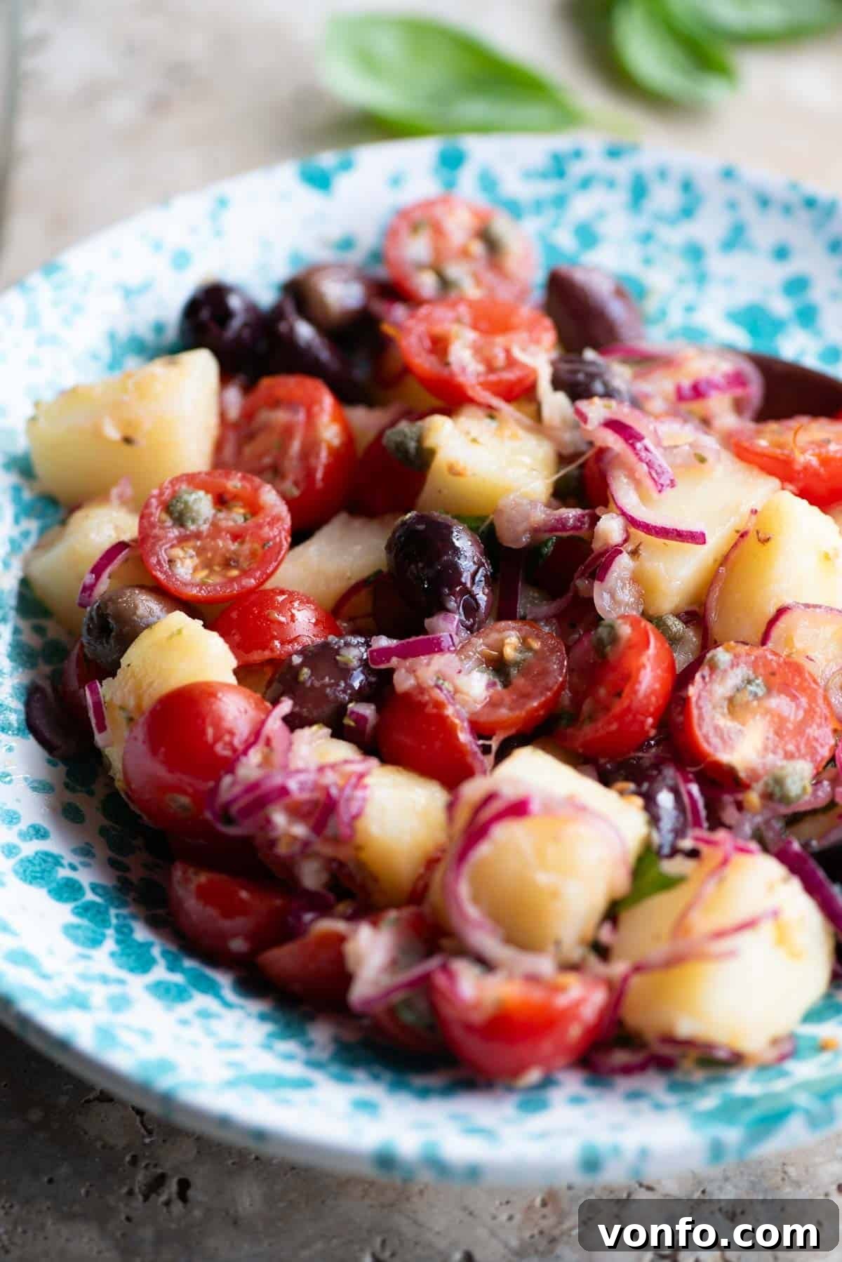 A close-up shot of a vibrant Sicilian potato salad, Insalata Pantesca, served in a blue and white patterned bowl, highlighting the textures of potatoes, olives, tomatoes, and herbs.