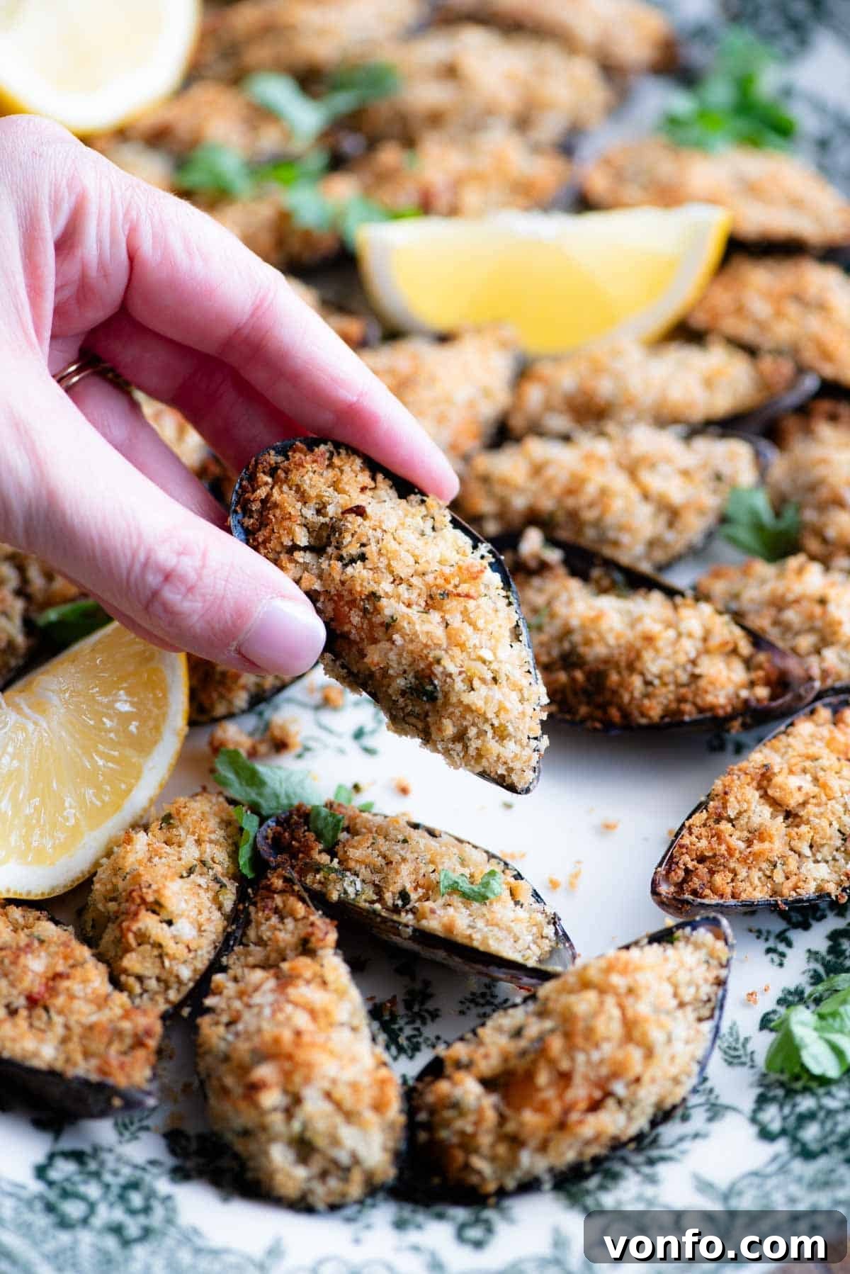 A close-up shot of a hand holding a single perfectly baked Italian stuffed mussel over a blurred serving plate, highlighting its golden, crispy topping.