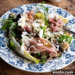 A close up cropped image of an eggplant and farro salad on a blue plate with prosciutto and pine nuts.
