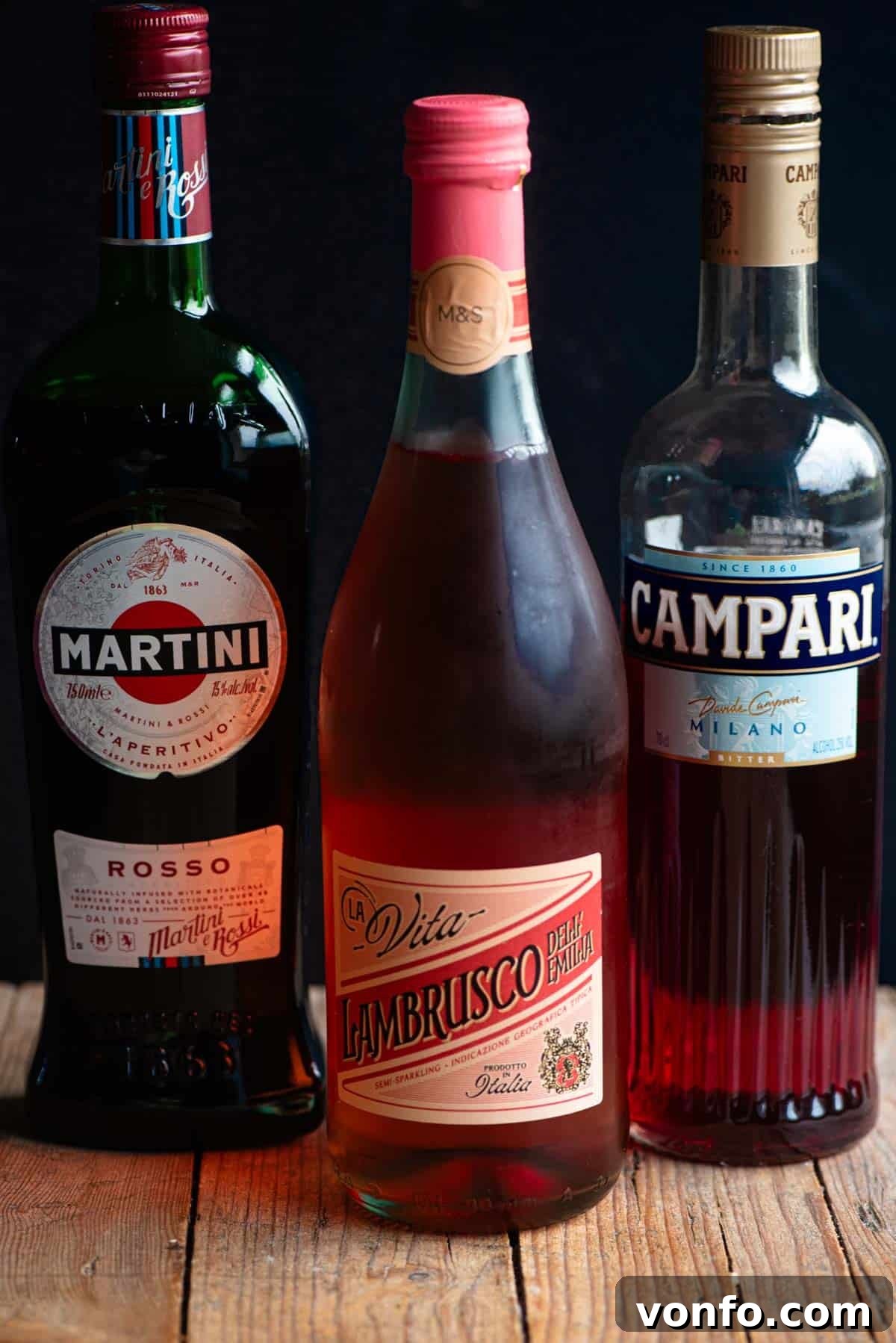 A bottle of Martini Rosso, Lambrusco, and Campari sitting on a wooden table with a black background, essential ingredients for the Sbagliato Rosso.