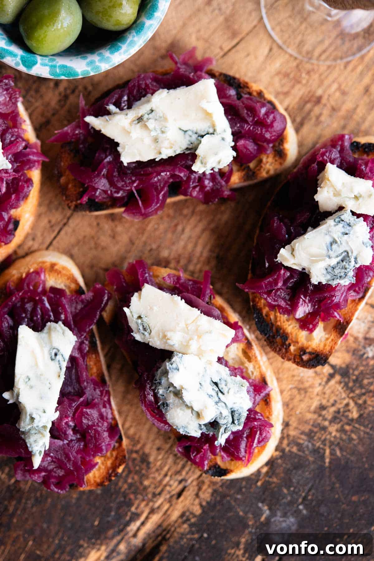 An overhead shot of red onion crostini topped with generous amounts of crumbled gorgonzola cheese on an elegant wooden serving board, ready for a festive gathering or party.