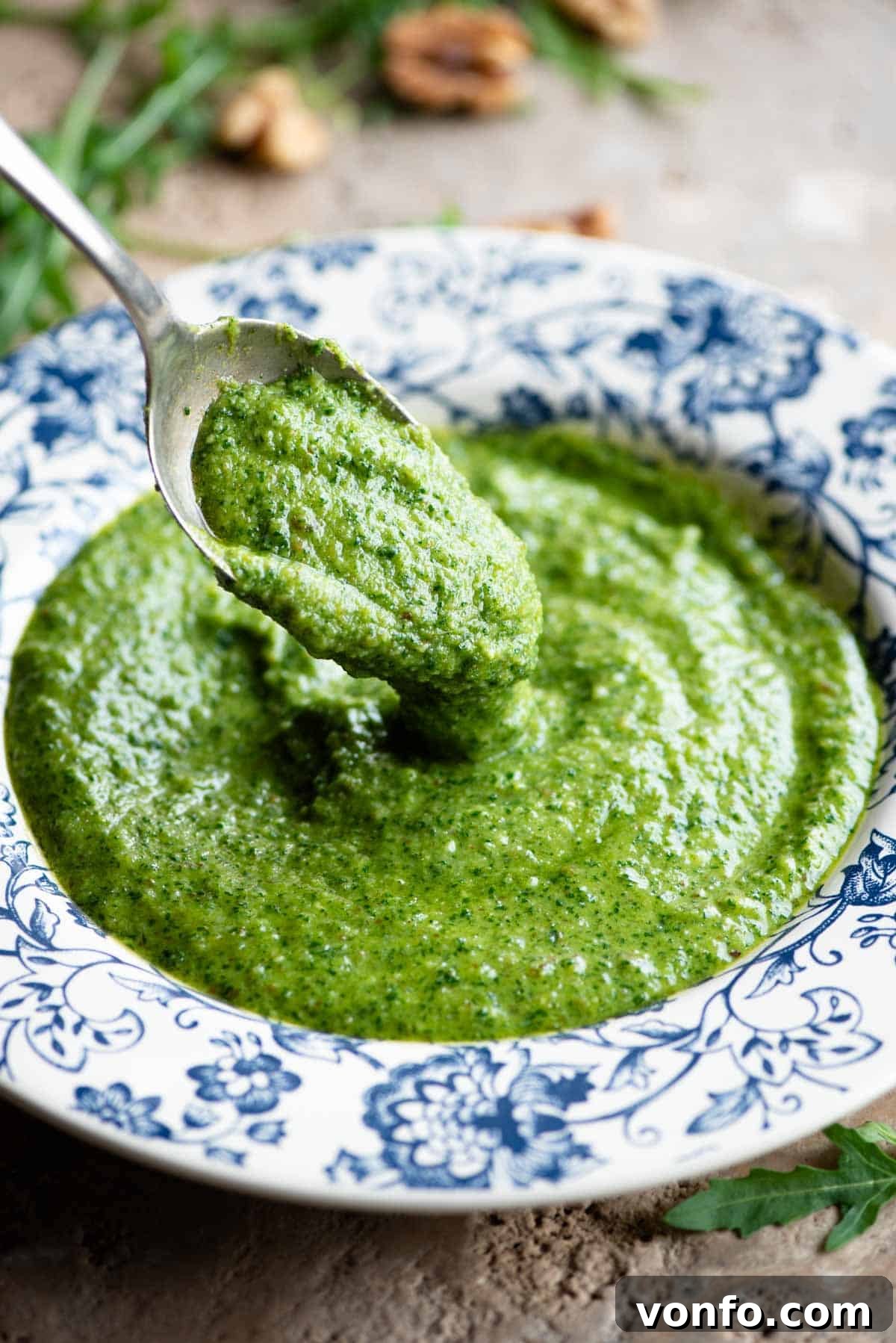 A close-up view of a spoon scooping up fresh, green arugula pesto from a patterned blue and white bowl, highlighting its rich texture.