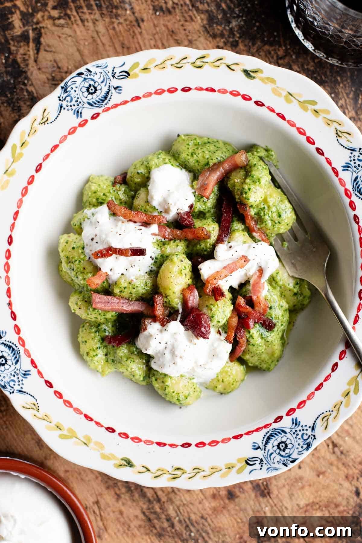 An overhead shot of arugula pesto gnocchi in a rustic bowl, generously topped with golden crispy pancetta and a dollop of creamy burrata, ready to be savored.