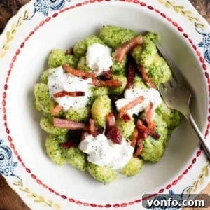 A close-up, cropped image of a bowl of arugula pesto gnocchi, beautifully garnished with crispy pancetta and a generous dollop of burrata, highlighting its rich textures and appealing presentation.