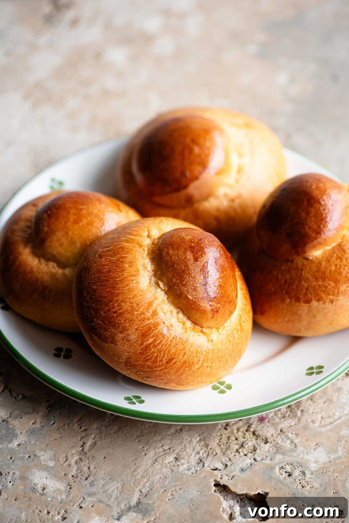 Four perfectly golden-brown Sicilian Brioche Buns (Brioche col Tuppo) arranged on an elegant plate, resting on a rustic travertine surface, inviting a taste of Italy.