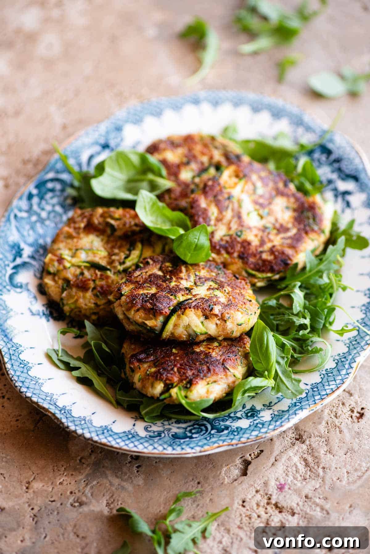 Five golden zucchini patties arranged on a bed of fresh arugula, served on a beautiful blue and white vintage-style platter resting on a travertine surface.