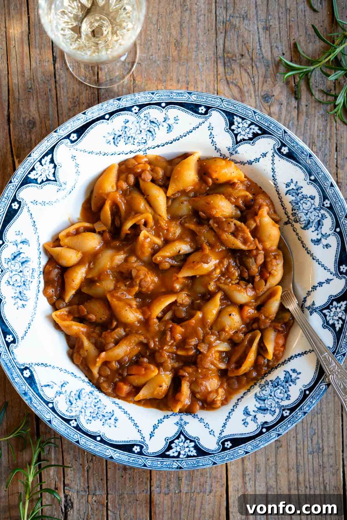 An overhead shot of a generous serving of Pasta e Lenticchie in a beautiful blue vintage-style bowl, garnished with fresh herbs, resting on a rustic wooden background. A glass of crisp white wine sits elegantly to the side, enhancing the inviting scene.