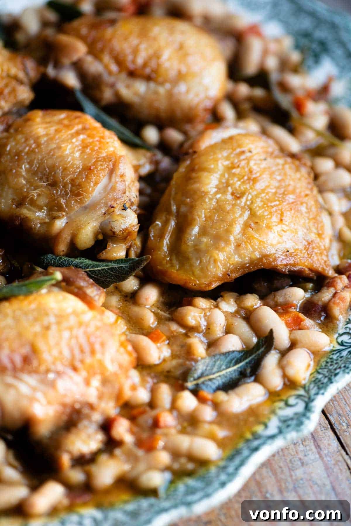 A close-up of a perfectly braised chicken thigh served on a rustic plate, nestled amongst creamy cannellini beans and fresh sage leaves, ready to be enjoyed.