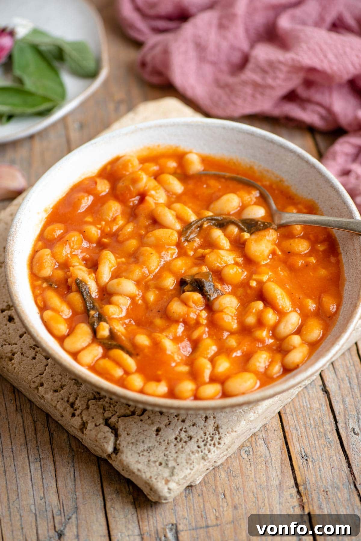 Tuscan White Beans in Rich Tomato Sauce 2 A bowl of creamy cannellini beans in a rich tomato sauce, garnished with fresh sage leaves, resting on a rustic wooden surface.