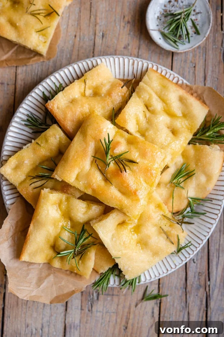 An overhead shot of golden-brown rosemary focaccia on a plate, garnished with fresh rosemary sprigs and sea salt flakes. The bread is cut into squares, showcasing its airy interior.