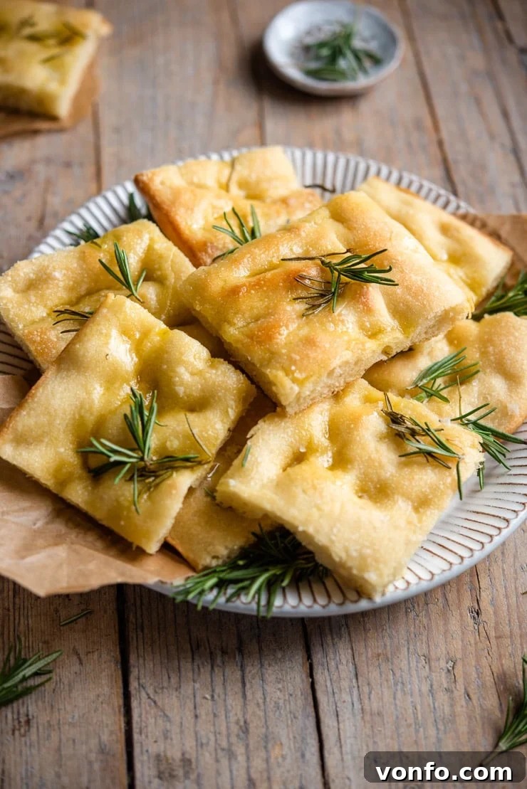 A close-up side view of a large, freshly baked rosemary focaccia, cut into individual squares and arranged on a rustic serving plate, highlighting its soft interior and golden crust.