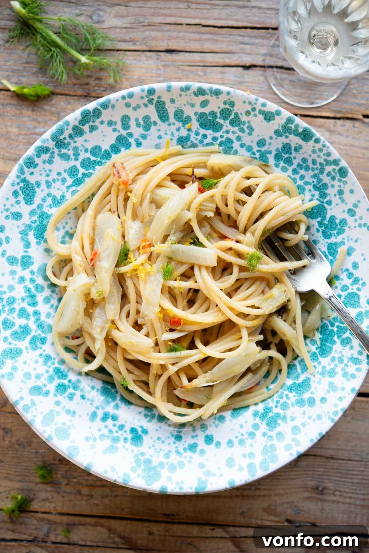 An overhead shot of roasted fennel pasta in a blue spotted bowl sitting on a rustic wooden surface, garnished with fresh fennel fronds.