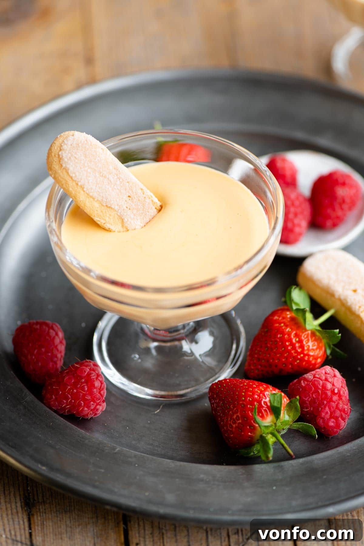 A small glass bowl filled with golden Zabaglione, with a Savoiardi cookie elegantly dipped into its creamy texture.