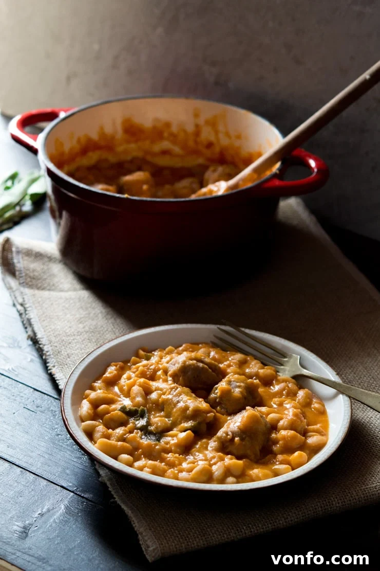 Tuscan bean stew with sausages on a plate with a fork at the side and a pot in the background, showcasing its rustic appeal.