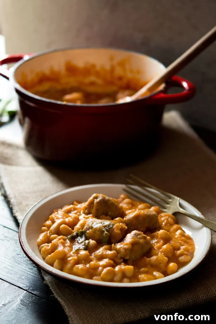 A close-up of a serving of Tuscan bean stew with sausages, showing the rich texture and vibrant colors, with a fork and the cooking pot blurred in the background.