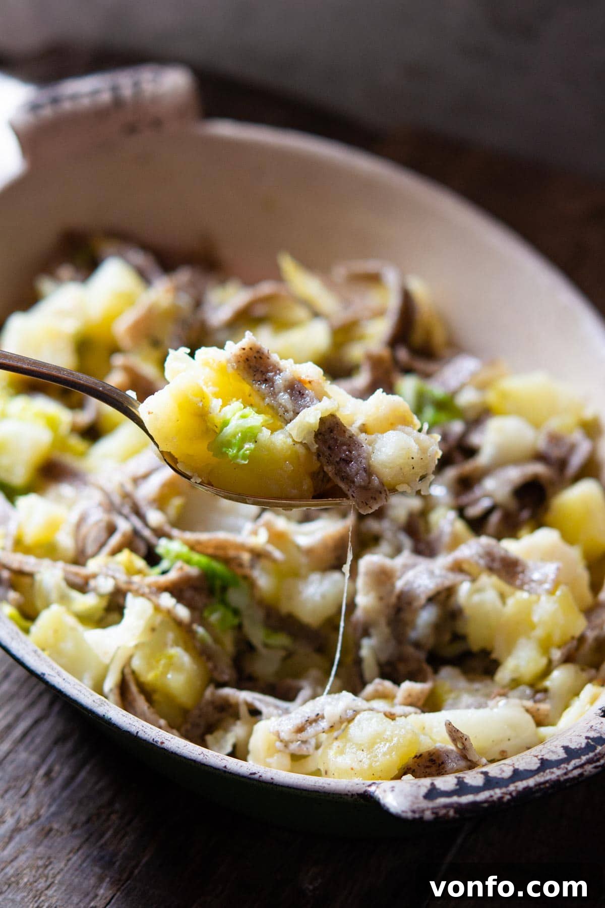 A close-up shot of a generous spoonful of Pizzoccheri pasta, prominently featuring the dark buckwheat noodles, tender golden potatoes, vibrant green cabbage, and rich, melted cheese, all glistening with aromatic garlic butter.