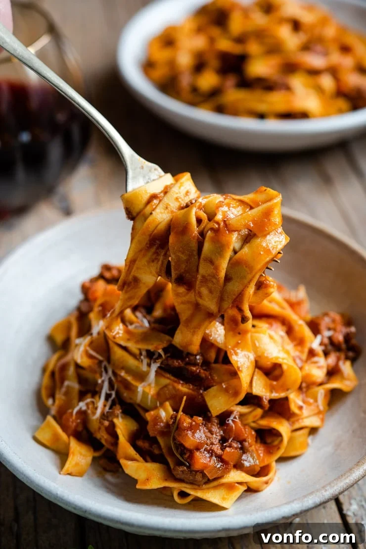 Authentic Italian Beef Ragu 2 A fork lifts a generous, glistening portion of rich Italian beef ragu intertwined with fresh tagliatelle pasta from a deep white bowl, showcasing the sauce's thick and luxurious texture.
