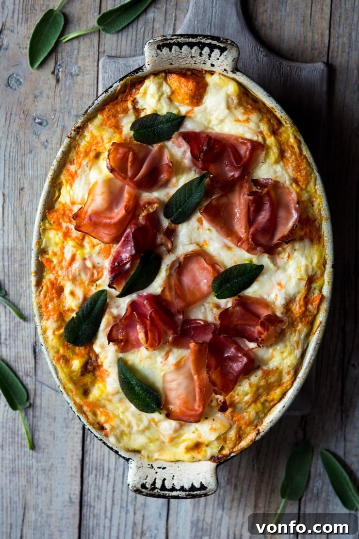 An overhead shot of butternut squash lasagna in an oval baking dish topped with prosciutto and sage leaves, showcasing its rich golden-brown top