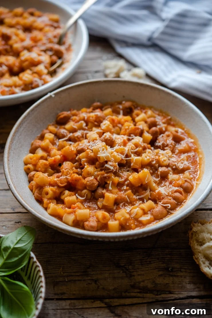 A beautifully presented bowl of pasta fagioli soup in a rustic bowl, garnished with fresh basil, accompanied by a side of crusty bread and a spoon on a wooden surface.