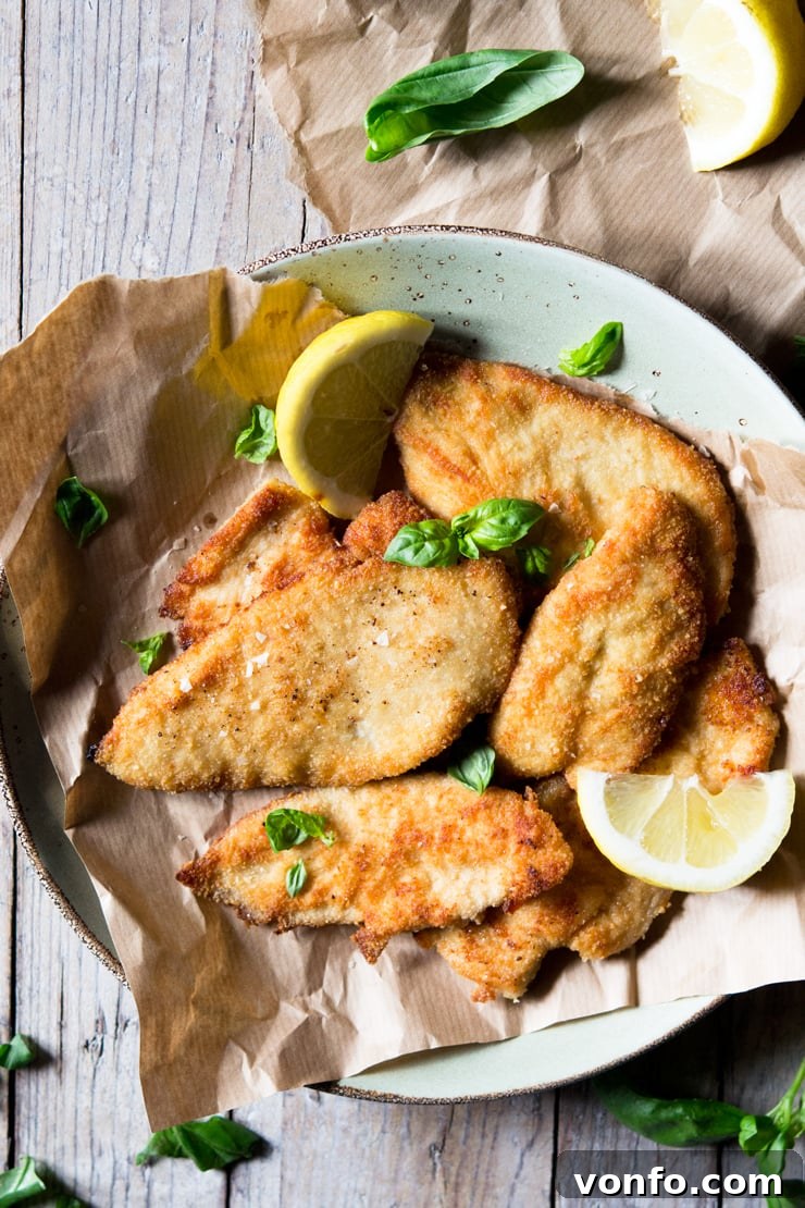 Crispy Breaded Chicken Cutlets with Lemon and Basil An overhead shot of golden-brown, crispy breaded chicken cutlets on a white plate, elegantly garnished with fresh basil leaves and lemon wedges, ready to be served.