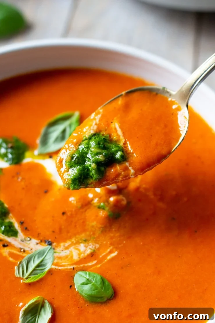 Roasted Red Pepper Soup. A delicious, thick and creamy soup made with roasted red peppers , sun dried tomatoes and topped with a pesto and creme fraiche swirl. A cozy and comforting soup for chilly nights. #peppersoup #soup A close-up shot of a spoon scooping up vibrant red roasted red pepper soup, beautifully topped with a swirl of green basil pesto and a creamy dollop.