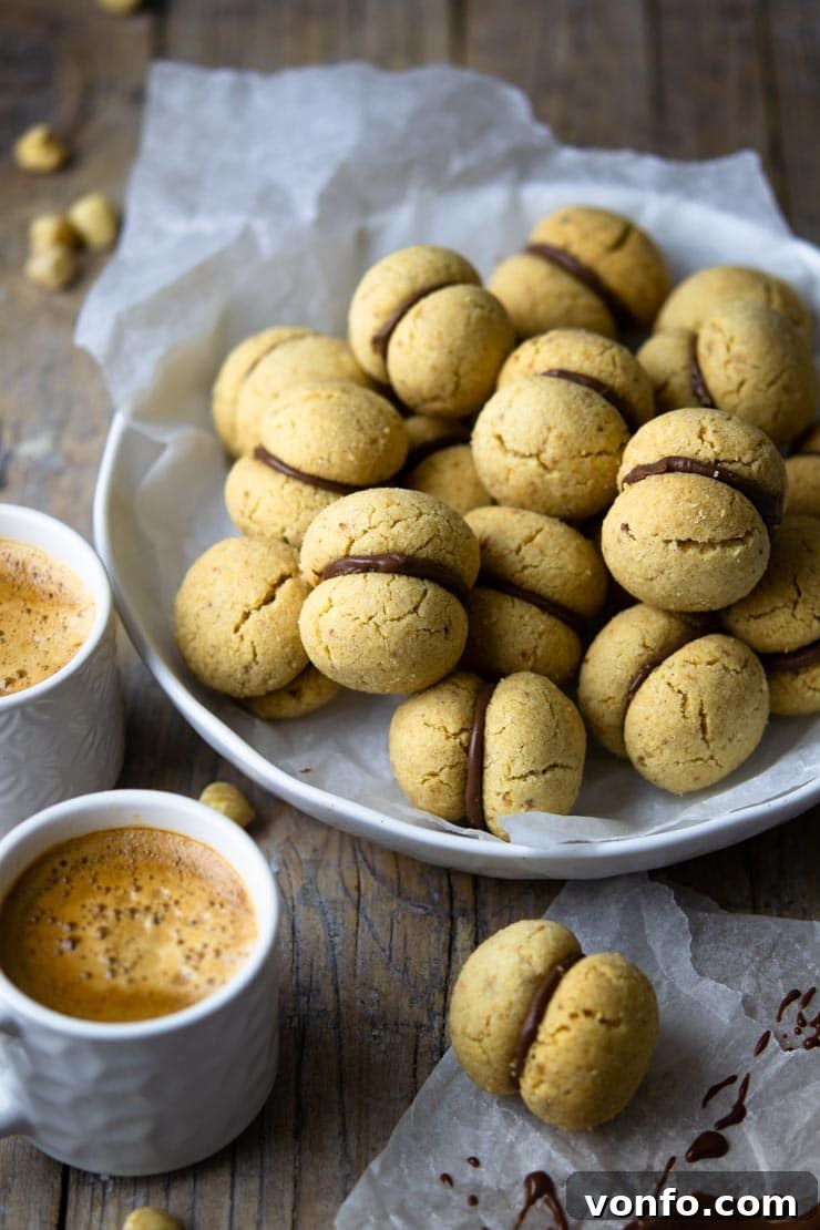 A close-up, beautifully styled shot of several Baci di Dama cookies artfully arranged in a rustic bowl, with two cups of coffee blurred in the background, inviting a moment of indulgence.