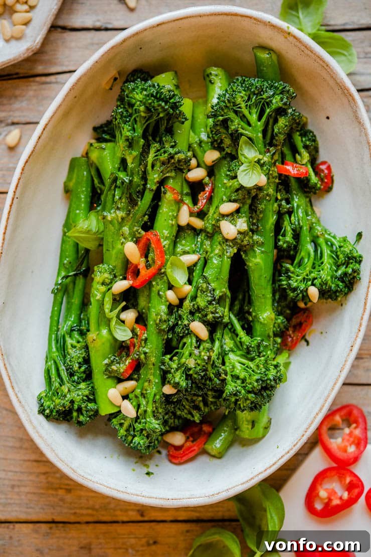 A close-up shot of vibrant green sautéed broccolini with visible pesto, pine nuts, and chili flakes in a serving bowl