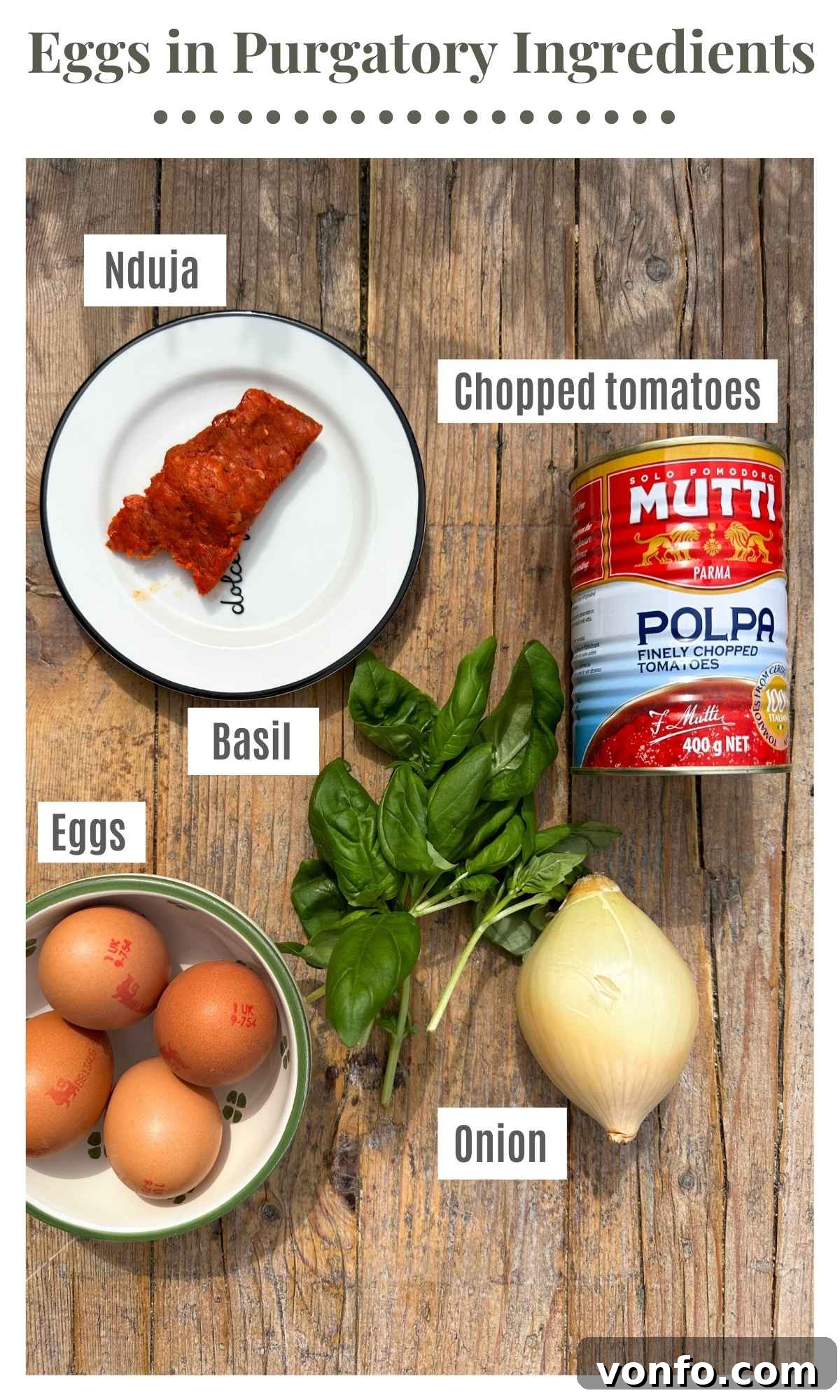 The ingredients to make spicy nduja eggs in purgatory on a rustic wooden surface. The ingredients are labelled: Nduja, chopped tomatoes, onion, basil, eggs.