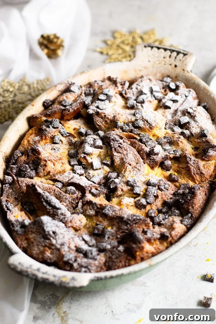Close-up of baked Panettone Bread Pudding. A close-up view of a baked Panettone Bread Pudding in a rustic oval baking dish, topped with melted chocolate chips and a dusting of powdered sugar.
