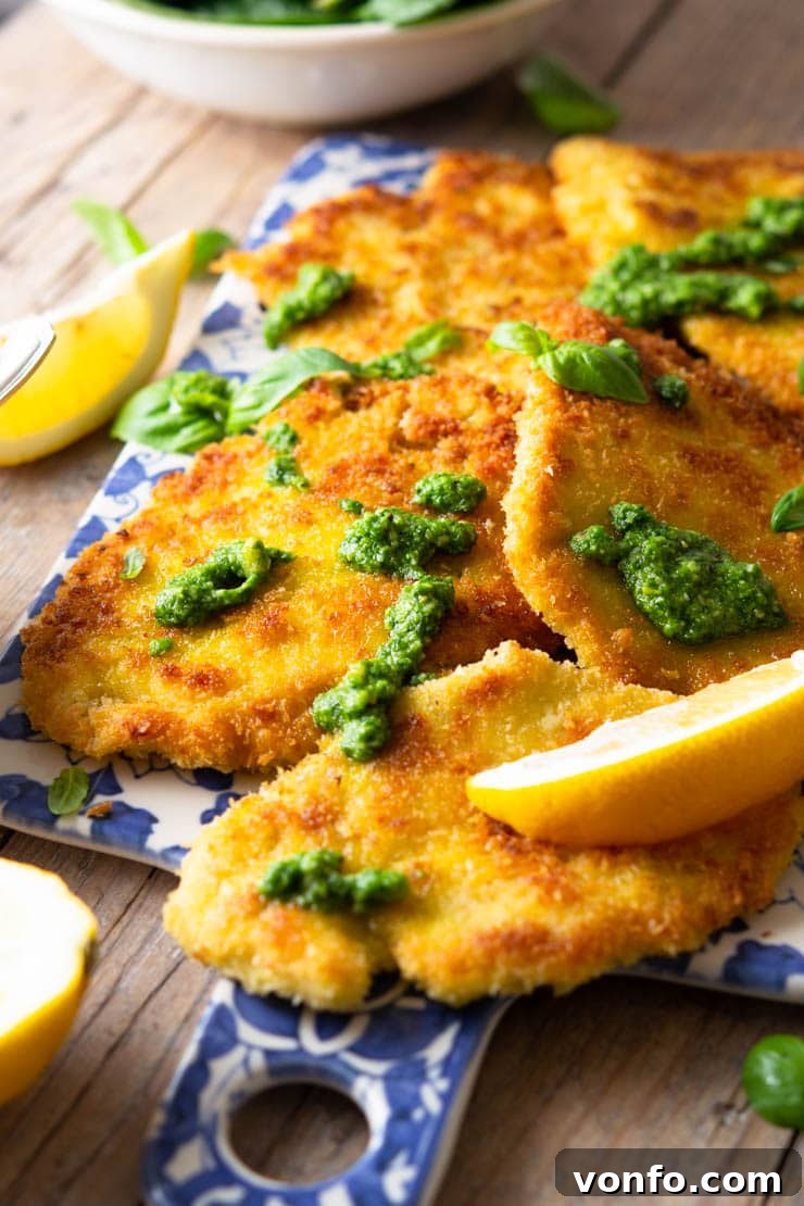 A close-up side view of golden-brown Italian chicken cutlets arranged on a blue serving board, artfully drizzled with fresh pesto and a sprinkle of lemon juice. The texture of the crispy breading is clearly visible.