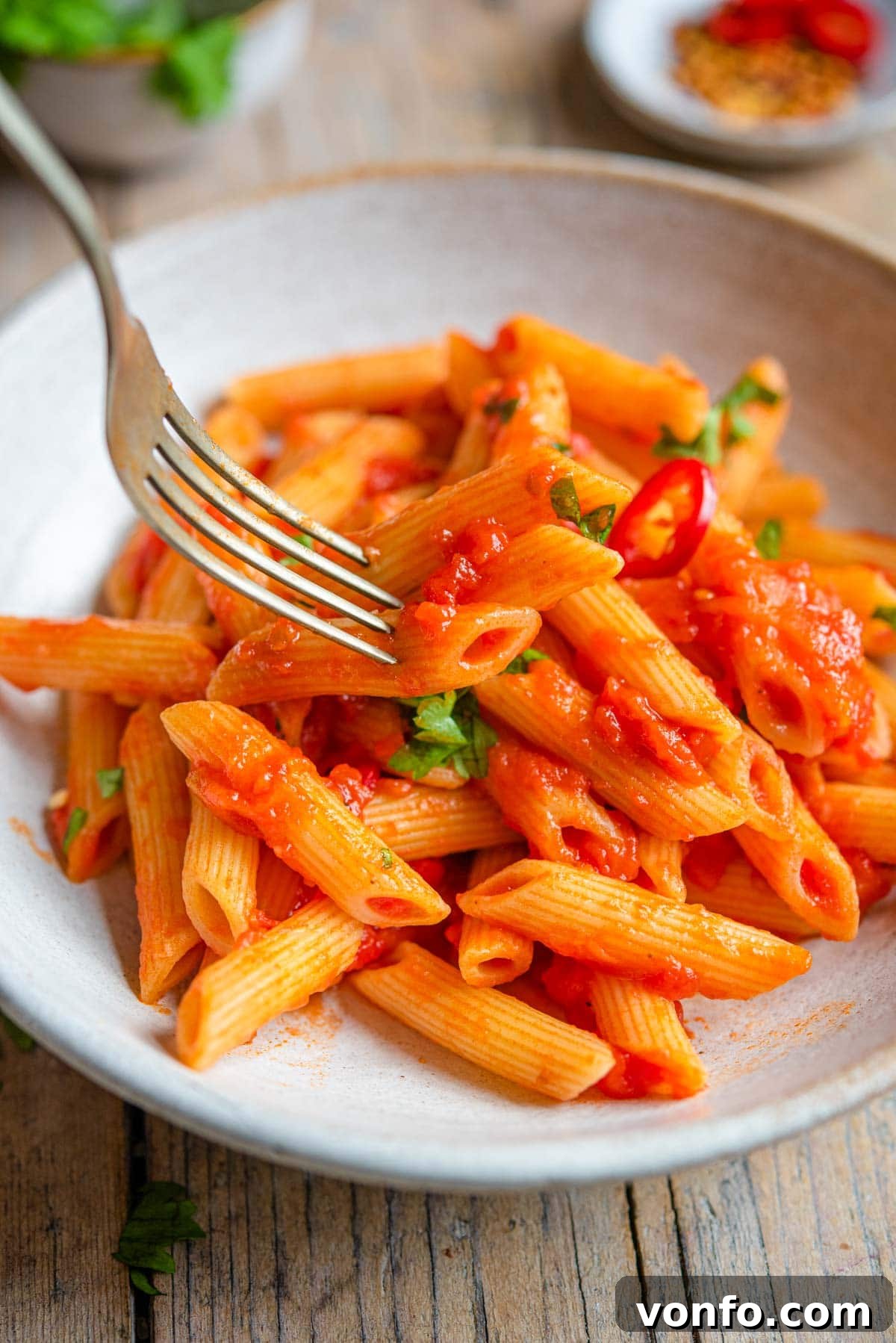 A close up of a fork picking up penne pasta from a bowl, showing the sauce clinging to the pasta
