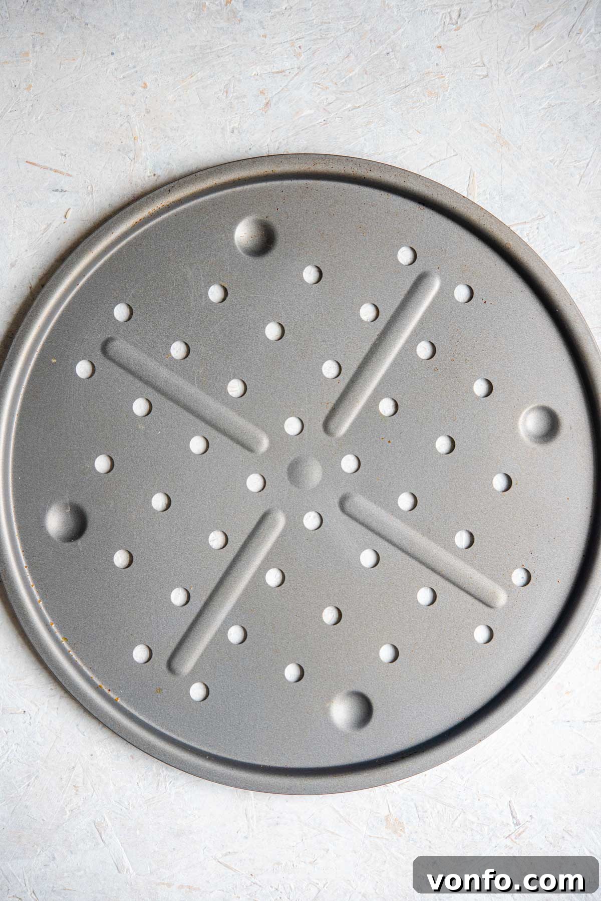 An overhead shot of a circular metal pizza tray with an array of small, evenly spaced holes across its surface, designed for superior heat distribution.