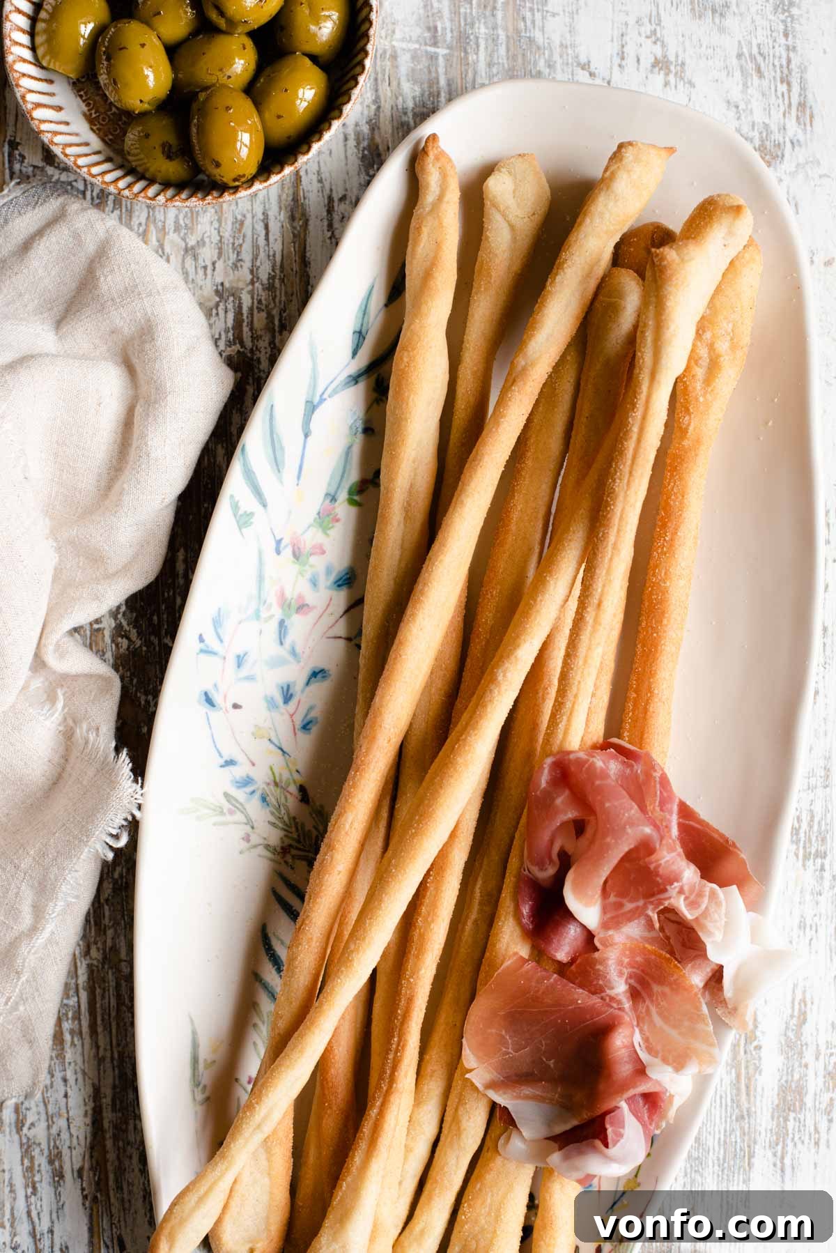 Grissini: Italy's Iconic Breadsticks 2 An overhead shot of Italian breadsticks on a serving plate with olives and prosciutto