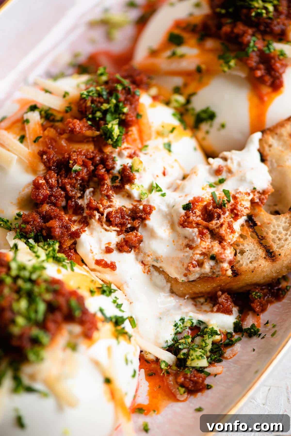 A close-up of a finished burrata cheese appetizer with crispy 'Nduja, gremolata, and grated Parmigiano Reggiano, served alongside slices of char-grilled ciabatta bread on a rustic wooden board.