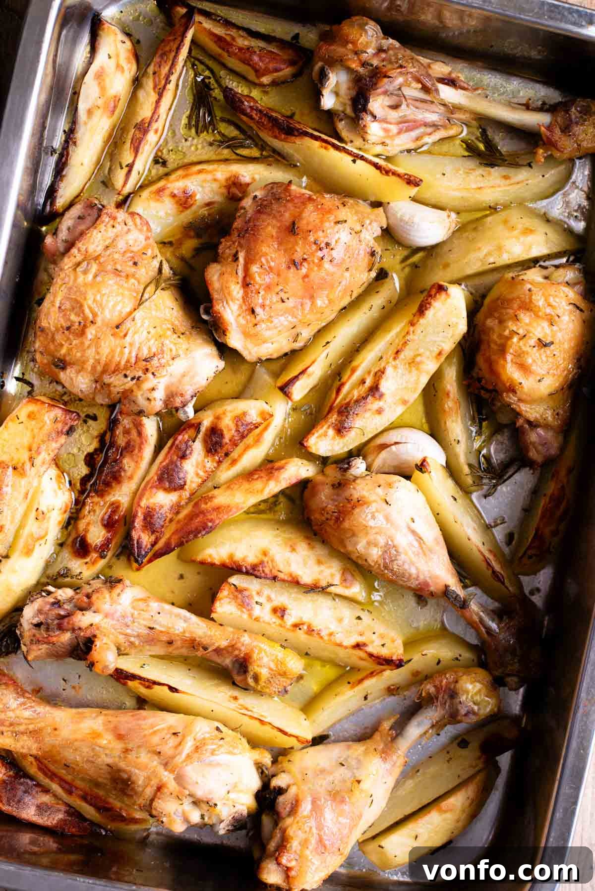 A close up overhead shot of golden-brown chicken and perfectly roasted potato wedges in a roasting tray, garnished with whole garlic cloves and fresh rosemary sprigs.